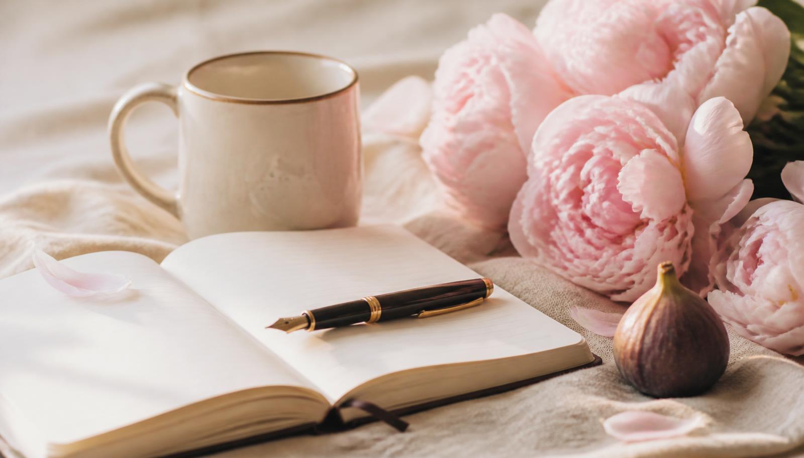 An open notebook, pen, mug and pink peonies on cream linen